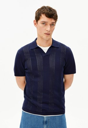 Junger Mann mit kurzen, lockigen braunen Haaren, der ein marineblaues Strickpolohemd mit vertikalen, strukturierten Streifen und hellblaue Jeans trägt und mit den Händen hinter dem Rücken steht.