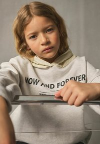 Jasnoszary sweatshirt z czarnym napisem, w tym "TERAZ I NA ZAWSZE", nakładany na kremowy hoodie. Ręce modela opierają się na przezroczystej powierzchni.