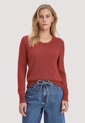 Frau trägt einen rostfarbenen Langarmpullover und hoch taillierte blaue Jeans mit gebundenem Kordelgürtel, steht vor einem schlichten Hintergrund.