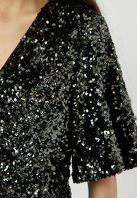 Tessuto nero con paillettes, design a scollo a V e maniche corte. Presenta paillettes riflettenti di diverse tonalità, creando una superficie con texture.