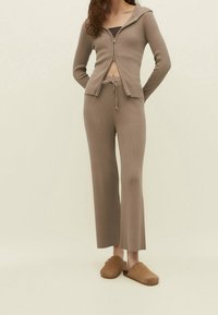 Chaqueta marrón de punto acanalado con cremallera y capucha, combinada con pantalones ajustados acanalados y zuecos marrones sin cordones, sobre un fondo neutro.