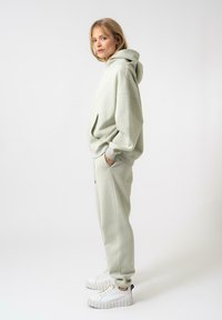 Felpa oversize verde chiaro con tasca a marsupio, abbinata a pantaloni della tuta coordinati e sneakers bianche chunky, caratterizzate da un tessuto testurizzato.
