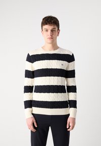 Dryžuota megzta pranašų palaidinė tamsiai mėlynos ir grietinėlės spalvų, turinti raštuotą apykaklę ir rankogalius, su siuvinėtų logotipu ant krūtinės.