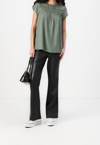 Frau in einem grünen ärmellosen Top mit Spitzenakzenten, schwarzen Lederhosen, weißen Turnschuhen und einer kleinen schwarzen Handtasche.