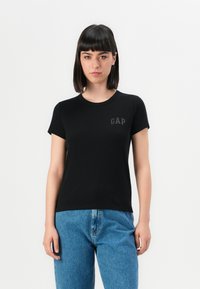 Donna con capelli scuri e lisci, che indossa una maglietta nera GAP e jeans blu, in piedi davanti a uno sfondo bianco semplice.