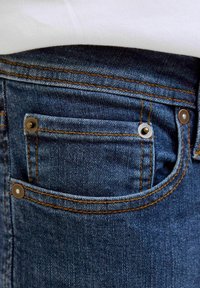 Vaqueros de mezclilla azul oscuro con costura de hilo dorado, que cuentan con un pequeño bolsillo delantero y dos remaches metálicos para refuerzo.