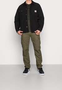 Veste en toile noire avec col marron, deux poches à l'avant, portée sur un pull vert texturé. Pantalons cargo olive et baskets noires.