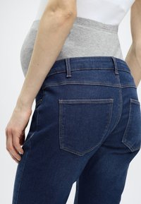 Σκούρο μπλε τζιν εγκυμοσύνης με ελαστική γκρι φάσα στην κοιλιά, διαθέτοντας πίσω τσέπες και ομαλή υφή τζιν. Εφαρμοστό σχέδιο που στενεύει στον αστράγαλο.