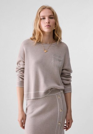 Giovane donna con capelli biondi che indossa un top a maniche lunghe color taupe e una gonna coordinata con dettagli a bottoni e una collana d'oro.