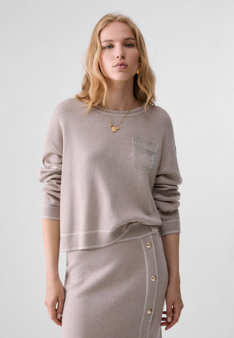 Jeune femme aux cheveux blonds portant un haut taupe à manches longues et une jupe assortie avec des détails à boutons et un collier doré.