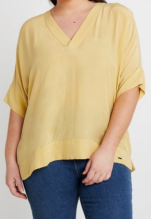 Persoon die een loszittende gele V-hals top met korte mouwen draagt, gecombineerd met een blauwe jeans, waarbij het bovenlichaam van nek tot heupen zichtbaar is.