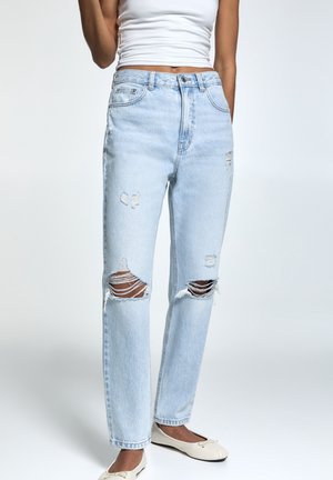 Woman wearing light blue ripped jeans paired with white tank top and beige ballet flats, standing against plain background.