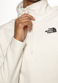 Sudadera de forro polar blanca con cuello alto, media cremallera y logotipo negro bordado en el pecho. Textura suave y mangas ajustadas.