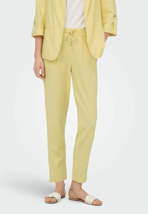 Kvinde iført lysegule bukser med snor, matchende blazer med opkædte ærmer, hvide sandaler og hvid top, stående mod ensfarvet baggrund.