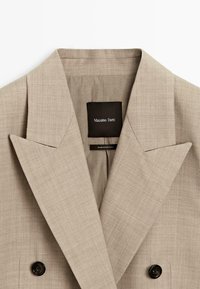 Beige skräddarsydd kavaj med breda slag, dubbla svarta knappar och en inre etikett som läser "Massimo Dutti" och "Tillverkat i Portugal."
