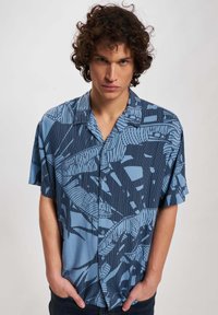 Camicia a maniche corte con bottoni di colore blu chiaro, decorata con un motivo geometrico audace di blu scuro. Realizzata in cotone, presenta una vestibilità rilassata e colletto.