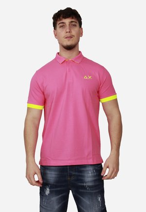 Uomo che indossa una polo rosa con bordi e simboli gialli sul petto, abbinata a jeans blu strappati, in piedi davanti a uno sfondo bianco.