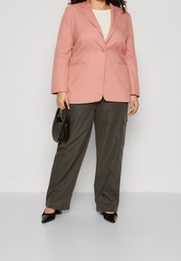Light pink blazer with a single button, worn over a cream top, paired with dark green, loose-fitting trousers and black pointed shoes.