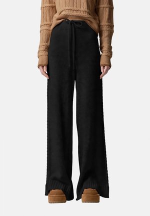 Persona con pantalones negros de pernera ancha con cintura ajustable, suéter marrón texturizado y zapatos marrones sobre un fondo liso.