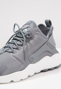 Baskets de sport en mesh gris avec semelle blanche, fermeture à lacets à l'avant et panneau de soutien du talon en plastique, photographiées sur un fond blanc.