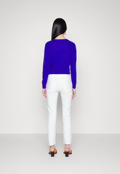 Élénk lila pulóver, slim fit fehér farmernadrág és narancssárga blokk sarkú szandálok. A sima textúrák és minimalista dizájn fokozzák az öltözéket.