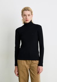 Person med kort blondt hår iført en tætsiddende sort turtleneck sweater og lysebrune fløjl bukser, stående mod en plain hvid baggrund.