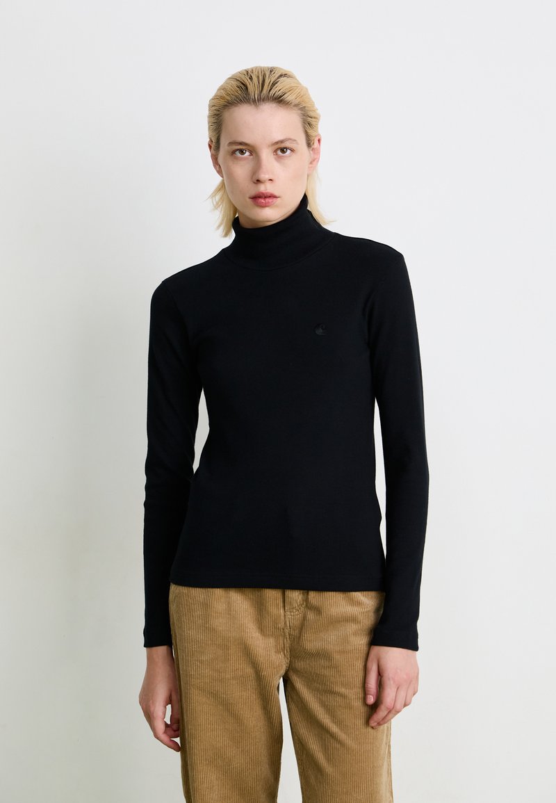 Person med kort blondt hår iført en tætsiddende sort turtleneck sweater og lysebrune fløjl bukser, stående mod en plain hvid baggrund.