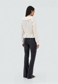 Blusa bianca e strutturata con collo a volant, vita cinched e maniche lunghe, abbinata a jeans a campana scuri. Vista posteriore mostrata.