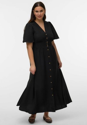 Femme portant une robe maxi noire à boutons avec des manches courtes bouffantes et des chaussures plates tissées, sur un fond blanc uni.