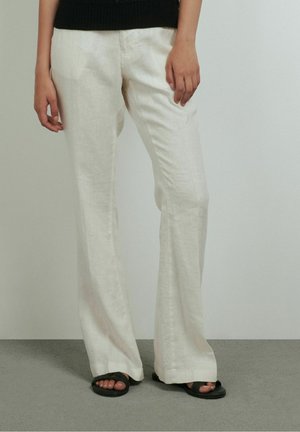 Persoon die losse witte linnen broek en zwarte sandalen draagt, staand op een grijze vloer tegen een lichtgrijze muur.