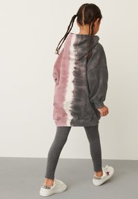 Bluza z kapturem w wzór tie-dye w kolorach różowym i szarym, z opuszczonymi ramionami i luźnym krojem, zestawiona z szarymi legginsami i białymi trampkami.