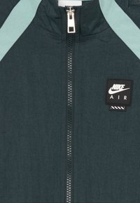 Nike Air jakna u tamno zelenoj boji s akcentima u svijetlo zelenoj. Ima puni zip i kvadratnu oznaku na prsima. Tekstuirana tkanina.