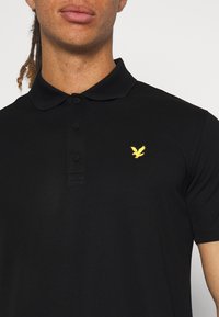 Mężczyzna w czarnej koszulce polo z trzema guzikami i małym żółtym logo ptaszka na lewej piersi.