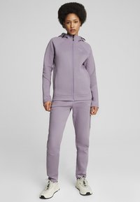 Chándal deportivo morado hecho de una tela suave y texturizada. Presenta una chaqueta con cremallera y capucha, y pantalones ajustados a juego.