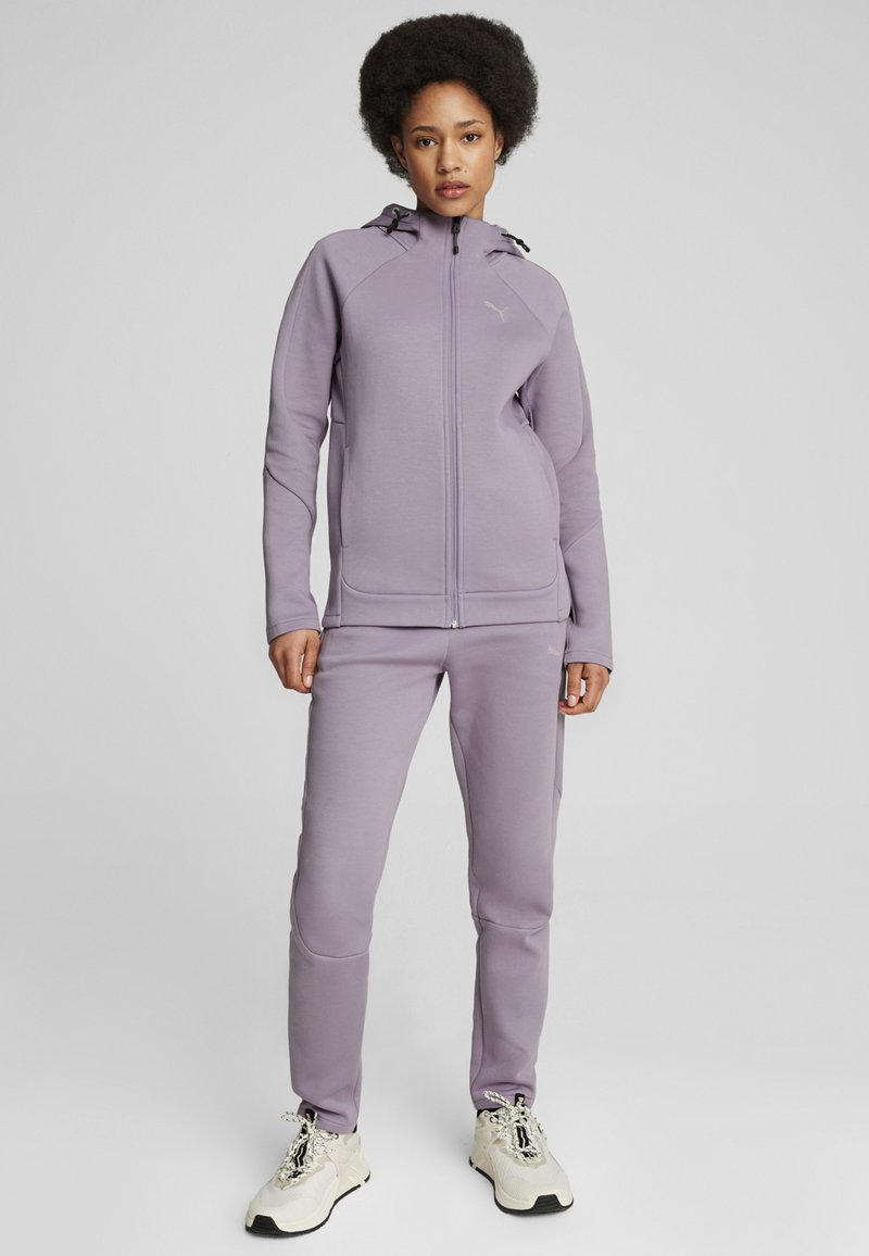 Chándal deportivo morado hecho de una tela suave y texturizada. Presenta una chaqueta con cremallera y capucha, y pantalones ajustados a juego.