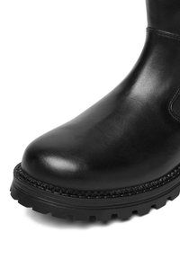 Czarny skórzany but z zaokrąglonym noskiem, ozdobnym przeszyciem i grubą, z bieżnikiem podeszwą na białym tle.