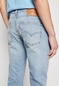 Osoba nosząca jasnoniebieskie dżinsy Levi's i włożoną niebieską koszulę, eksponująca tylną kieszeń oraz skórzaną metkę marki na pasku.