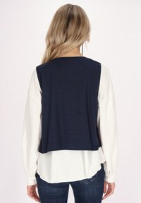 Marinblå ärmlös topp med texturerad yta, lager på lager över en vit långärmad blus, matchad med mörka denimjeans.