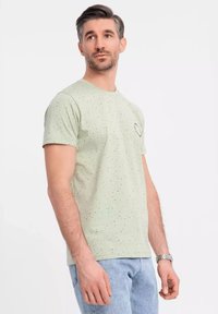 Lichtgroen T-shirt met korte mouwen, voorzien van zwarte spikkels en een kleine grafische afbeelding op de linkerkant van de borst. Zachte katoenkwaliteit, ontspannen pasvorm.