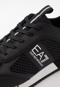 Scarpa sportiva nera con materiali in rete e sintetici lisci. Presenta lacci tonalizzati, una patch con logo e una suola bianca con battistrada testurizzato.