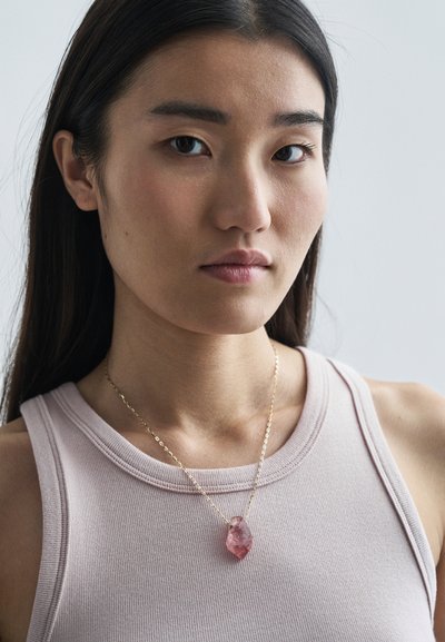 Zlatý reťazový náhrdelník s veľkým, nepravidelným ružovým kamenným príveskom, nosený cez svetloružový, ryhovaný top bez rukávov. Hladký povrch a prirodzené variácie.