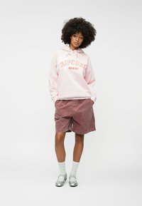 Sieviete ar afro matiem, tērpusies gaiši rozā Rip Curl Hawaii kapuce, mauve šortos, baltās zeķēs un sudrabainās Mary Jane apavās, stāv ar rokām kabatās.