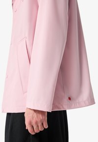 Mann trägt eine hellrosa, knöpfbare Jacke mit Seitentasche, sichtbarer linker Hand und schwarze Hose vor weißem Hintergrund.