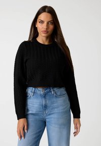 Maglione cropped nero a maglia con un motivo a zigzag testurizzato e polsini a coste, abbinato a jeans azzurri a vita alta. Bottoni dorati sulla spalla.