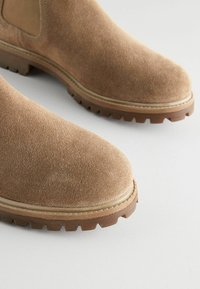 Bottines en daim marron avec un bout arrondi, une semelle en caoutchouc et des accents beige. La texture est douce, avec un petit talon et une semelle à crampons.