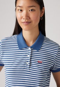 Μπλε και λευκή ριγέ πόλο μπλούζα με ομοιόμορφο μπλε γιακά, διαθέτει κόκκινο λογότυπο της Levi's. Έχει κοντά μανίκια και τρία κουμπιά στον γιακά.