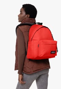Mochila Eastpak roja con un bolsillo delantero, correas de tela y detalles en negro. Lleva puesta sobre un chaleco acolchado marrón y leggings grises con textura.