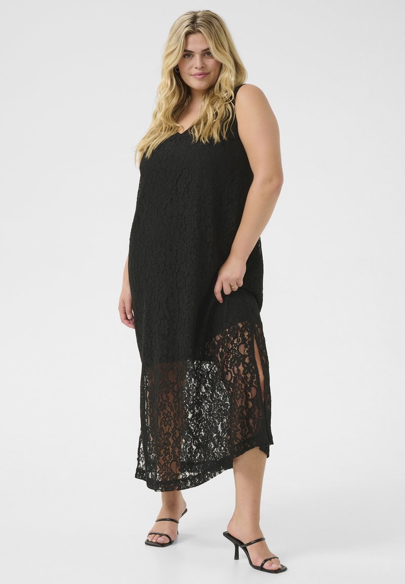 Une robe noire en dentelle avec un design sans manches, un look superposé, une partie inférieure translucide et un ourlet légèrement asymétrique. Accessoirisée avec des sandales.