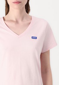 Šviesiai rožinė medvilninė marškinė su V formos iškirpte ir trumpomis rankovėmis, su mėlynu stačiakampiu "HUGO" logotipu kairiajame viršutiniame kampe.