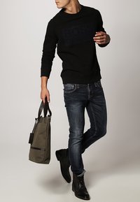 Sudadera negra con un logotipo texturizado, jeans azules, botas de cuero negras y una bolsa de lona verde olivo con una correa en contraste.